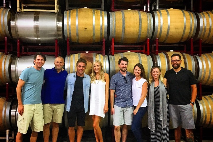 Group of eight people posing in front of stacked wine barrels in a winery.