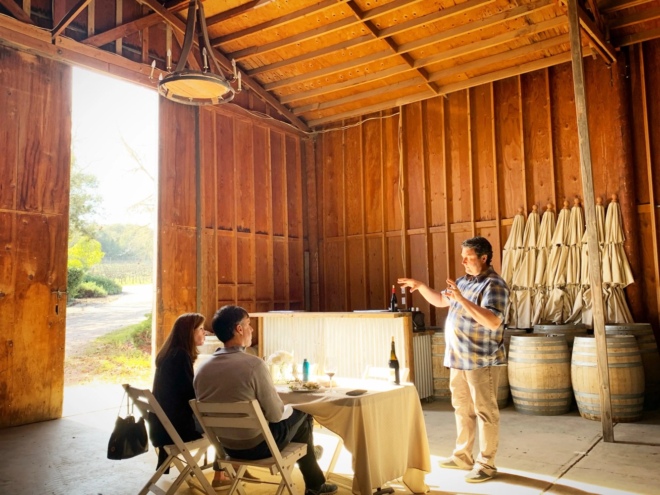 Person demonstrating wine to two seated individuals in a rustic wooden barn setting.