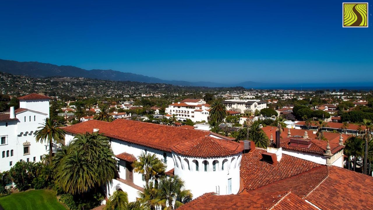 Cityscape with red-roofed buildings, palm trees, and mountains under a clear blue sky.