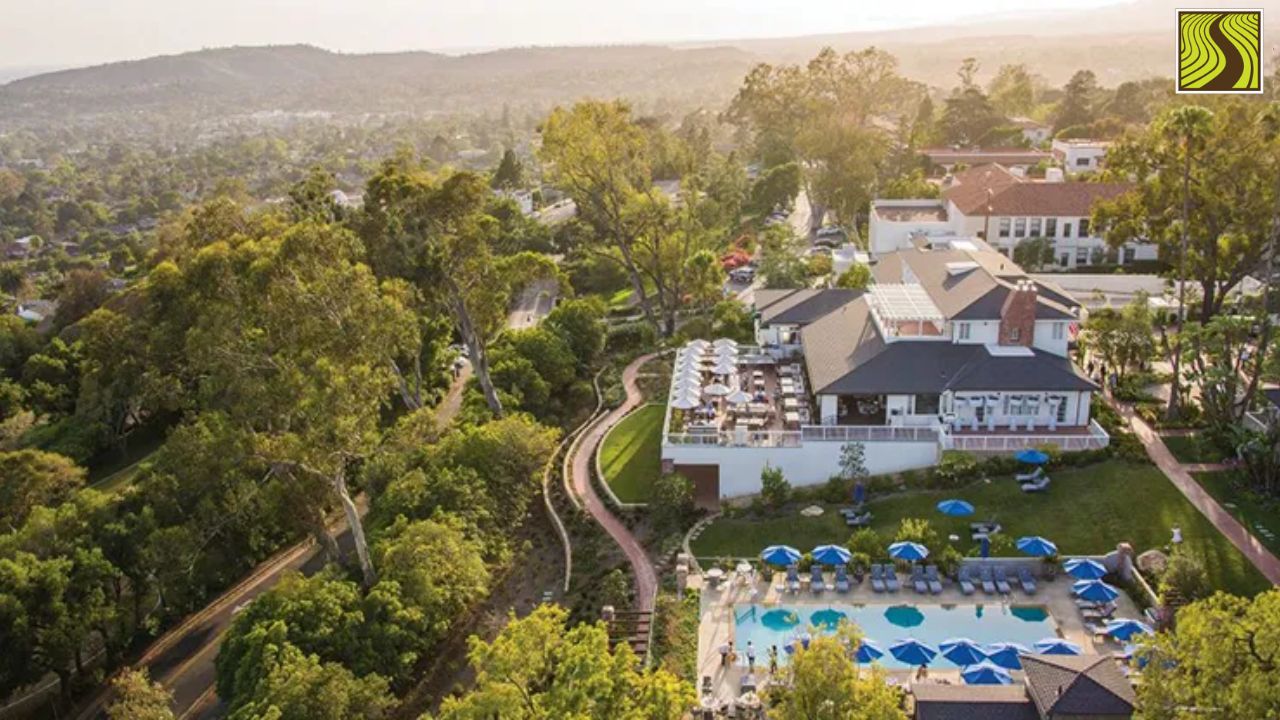Aerial view of a resort with a pool, umbrellas, and surrounding greenery on a sunny day.