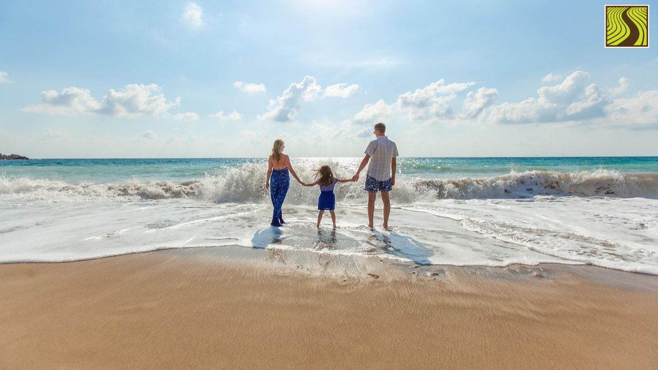 Family of three holding hands, standing on a sandy beach facing the ocean.