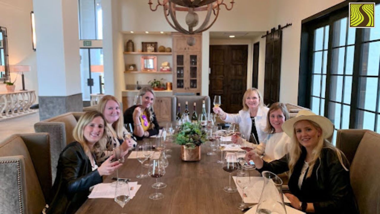 Six women smiling and holding wine glasses around a wooden table in an elegant room.