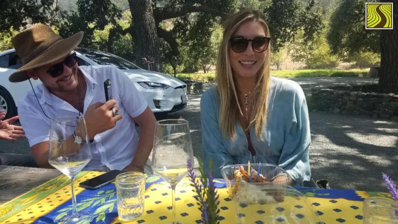 Two people sitting outside at a table with drinks, smiling under trees.