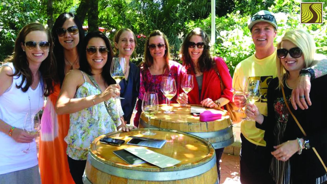 Group of people outdoors holding wine glasses, standing around barrel tables.