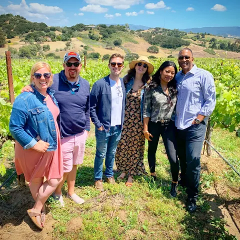 Group of six people posing in a sunny vineyard with hills in the background.