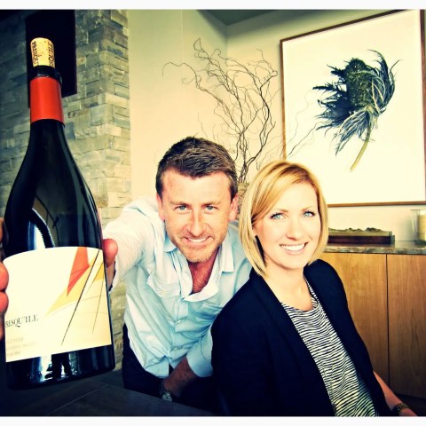 Person holding a wine bottle, another person smiling, with art on the wall behind them.