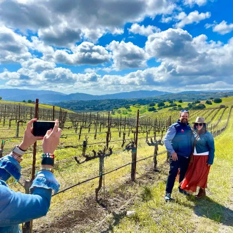 Person taking photo of a couple in a scenic vineyard under a partly cloudy sky.