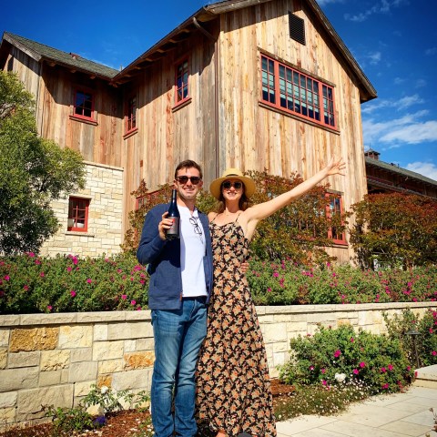 Couple posing with wine bottle, wearing casual outfits, in front of a rustic house and garden.