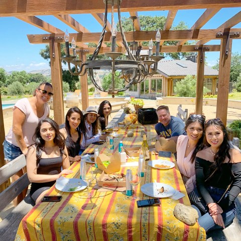 Group of people sitting at an outdoor table under a pergola, enjoying a meal.