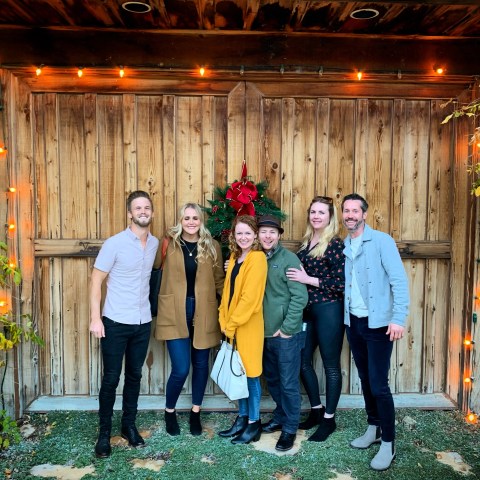 Six people posing together in front of a wooden barn door with festive lights and wreath.