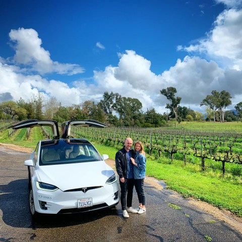 A couple stands beside a white car with open doors in a vineyard under a cloudy sky.
