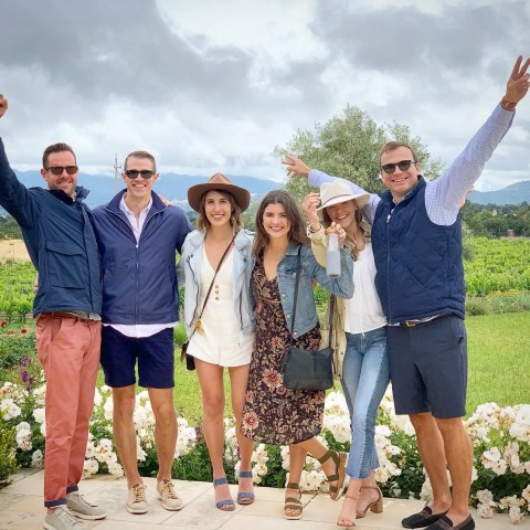 Group of six smiling people posing with arms raised in a scenic outdoor setting.
