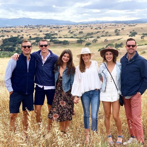 Group of six people standing in a field with hills and trees in the background, dressed casually.