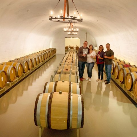 Four people standing in a wine cellar between rows of large barrels.