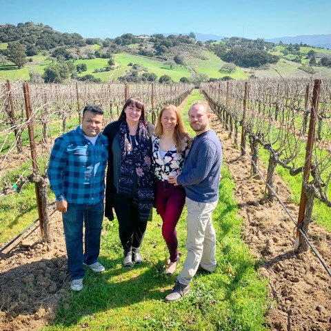 Four people standing in a vineyard with hills and blue sky in the background.
