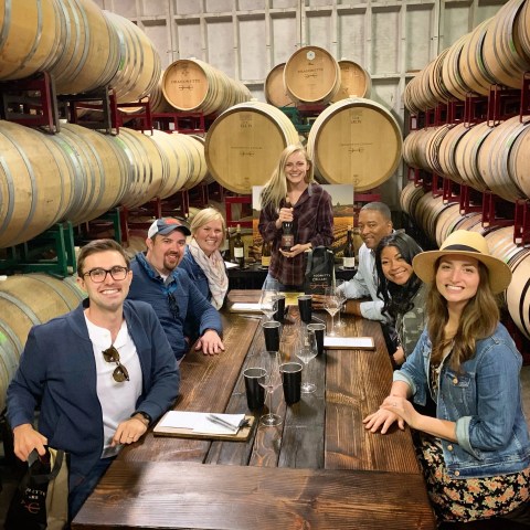 Group of people smiling at a table in a wine cellar with barrels.