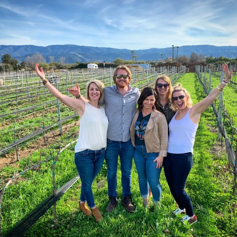 Group of five people smiling in a vineyard with mountains in the background.