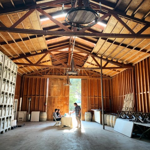 Two people at a table inside a wooden barn with stacked crates and barrels.