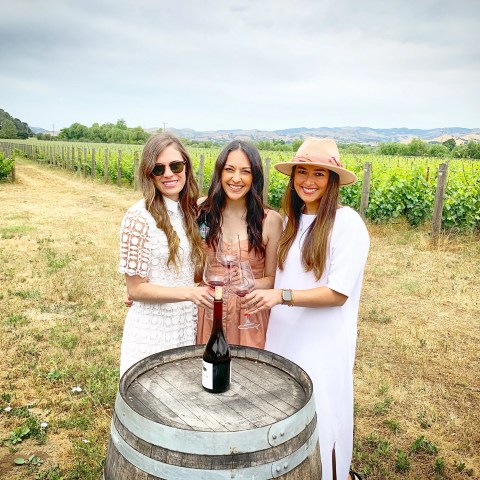 Three women standing by a wine barrel in a vineyard, holding glasses of wine.