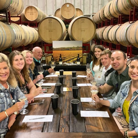 Group of people at a wine tasting in a barrel room, holding wine glasses and smiling at a table.