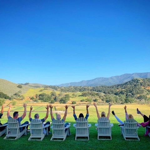 People sitting on chairs outdoors, raising arms towards blue sky, mountains in background.