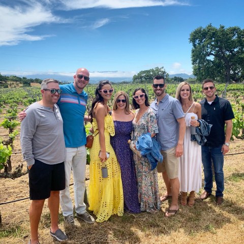 Group of seven people smiling in a vineyard under a blue sky.