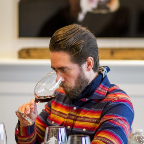 Man in colorful shirt sniffing red wine in a glass at a tasting table.