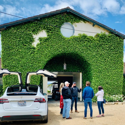 Group of people talking near a Tesla with open doors in front of an ivy-covered building.