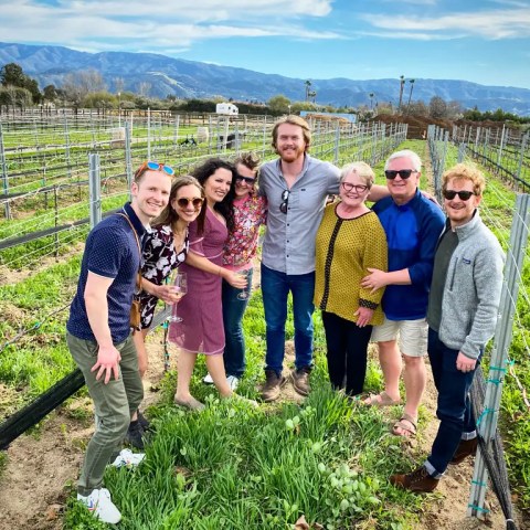 Group of seven people posing in a vineyard on a sunny day with mountains in the background.
