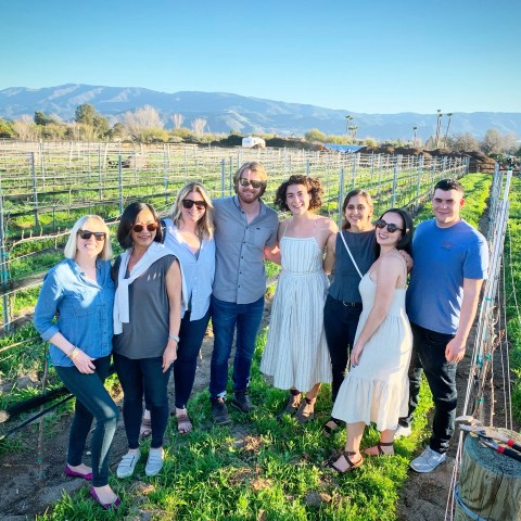 A group of eight people smiling in a vineyard with mountains in the background.
