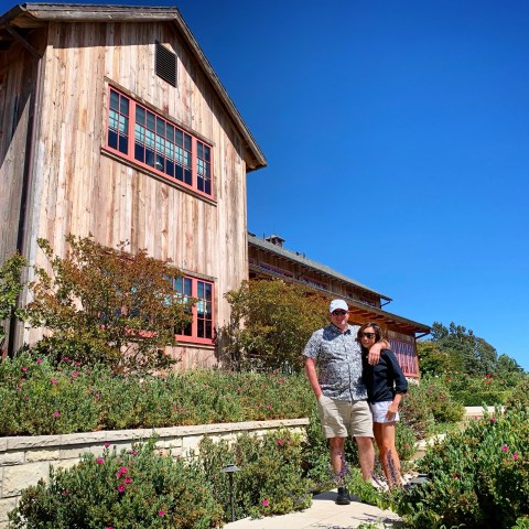 Two people standing in front of a rustic wooden house with red-framed windows and garden, under a clear blue sky.