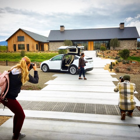 Photographers capturing a man walking by a white car with open doors in front of a modern house.