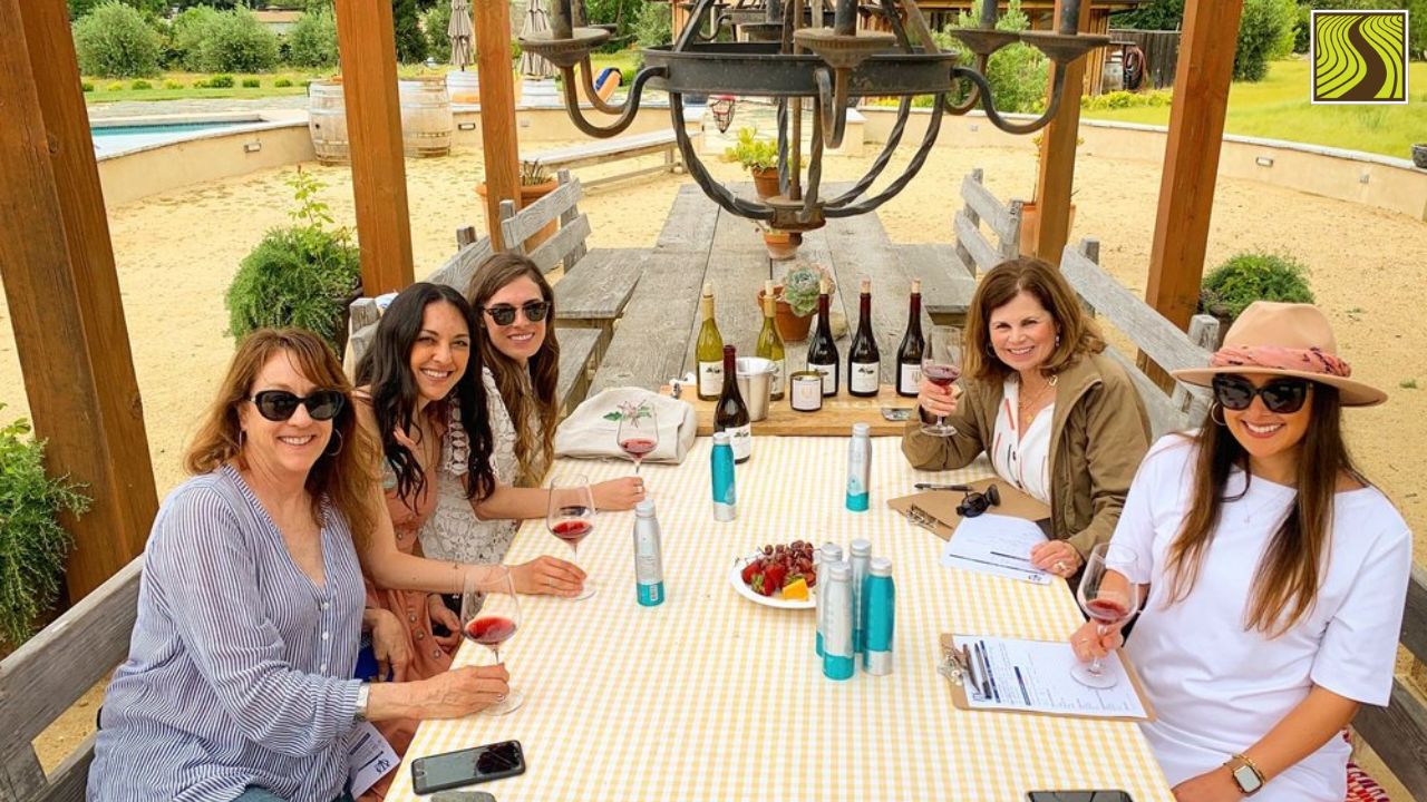 Five women seated at a picnic table with wine glasses and bottles outdoors.