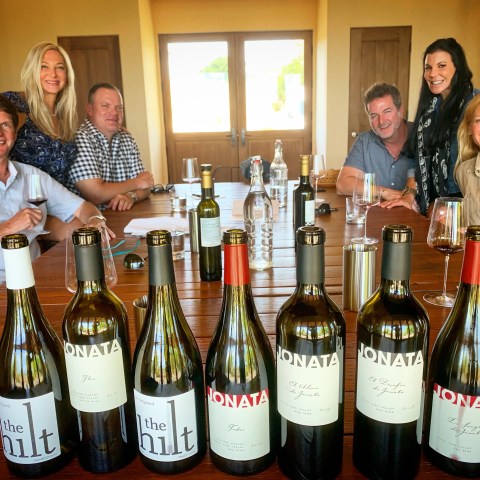 Six people sitting at a table with bottles of wine and glasses.