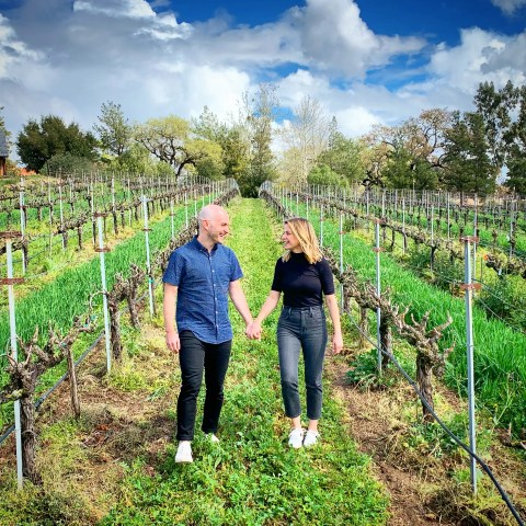 Couple holding hands, walking through a vineyard under a cloudy sky.
