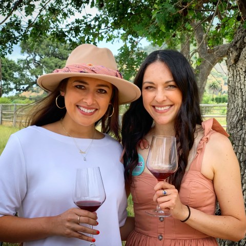 Two women smiling, holding wine glasses in a scenic outdoor setting with trees.