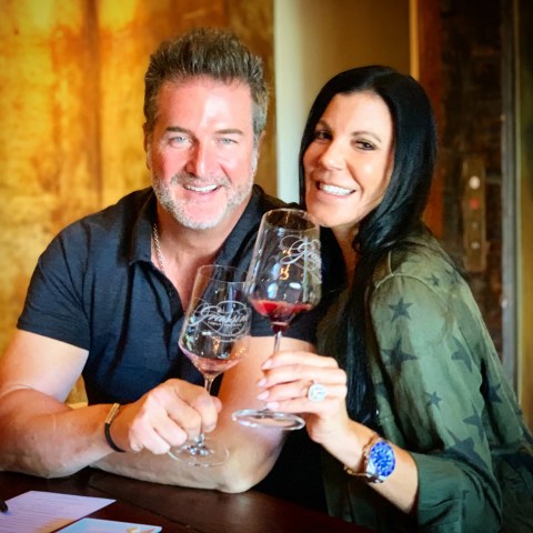 A smiling couple toasting with wine glasses at a table indoors.