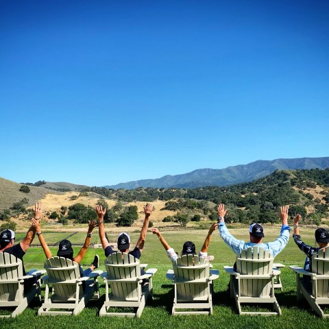 Six people in chairs raising arms, facing hills and clear sky.