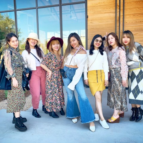 Seven women in stylish outfits pose together outside a modern building with glass windows.