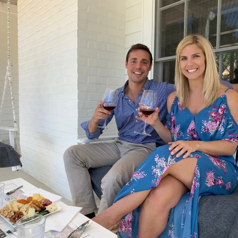 Couple sitting on porch, holding wine glasses, with a cheese platter and wine bottle on the table.