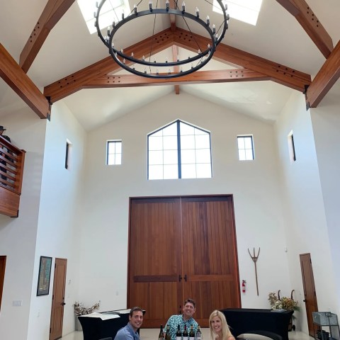 Three people seated at a long table in a high-ceilinged room with wooden beams and a circular chandelier.