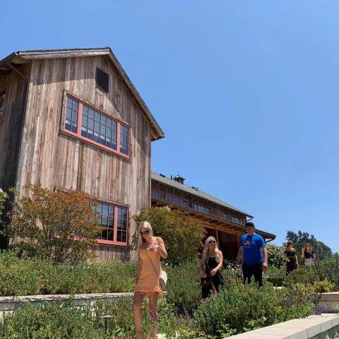 Group of people walking past a rustic wooden house in a garden under a clear blue sky.