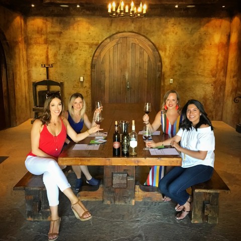 Four women sitting at a rustic table in a cellar-like room, holding wine glasses and smiling.
