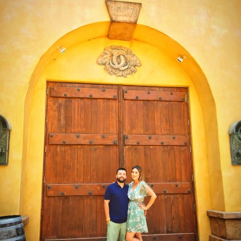 A couple stands in front of large arched wooden doors, with a yellow wall and Roman numerals above.