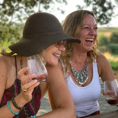 Two women laughing and holding wine glasses at an outdoor table.