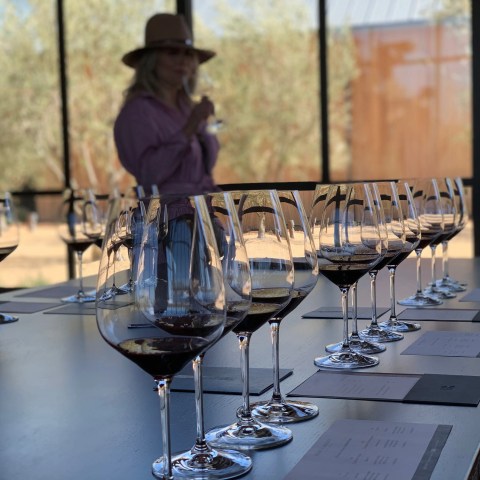 Row of wine glasses on a table; woman in hat tasting wine in background.