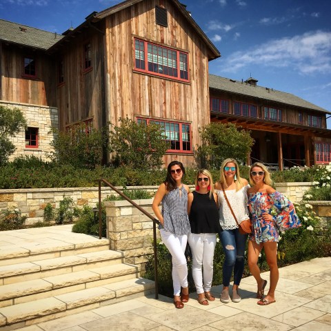 Four women posing on a sunny day in front of a large, rustic wooden house with a stone walkway.