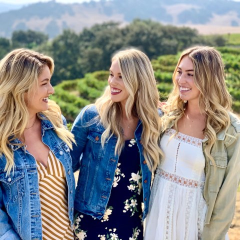 Three women in casual dresses and jackets smiling at each other in a vineyard with hills in the background.