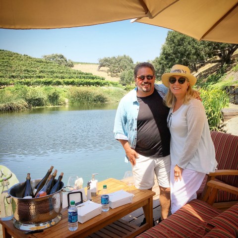 Two people smiling by a lakeside with picnic table and wine bottles under an umbrella.