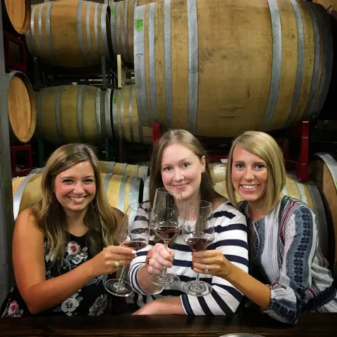 Three women in a winery toasting with wine glasses in front of barrels.